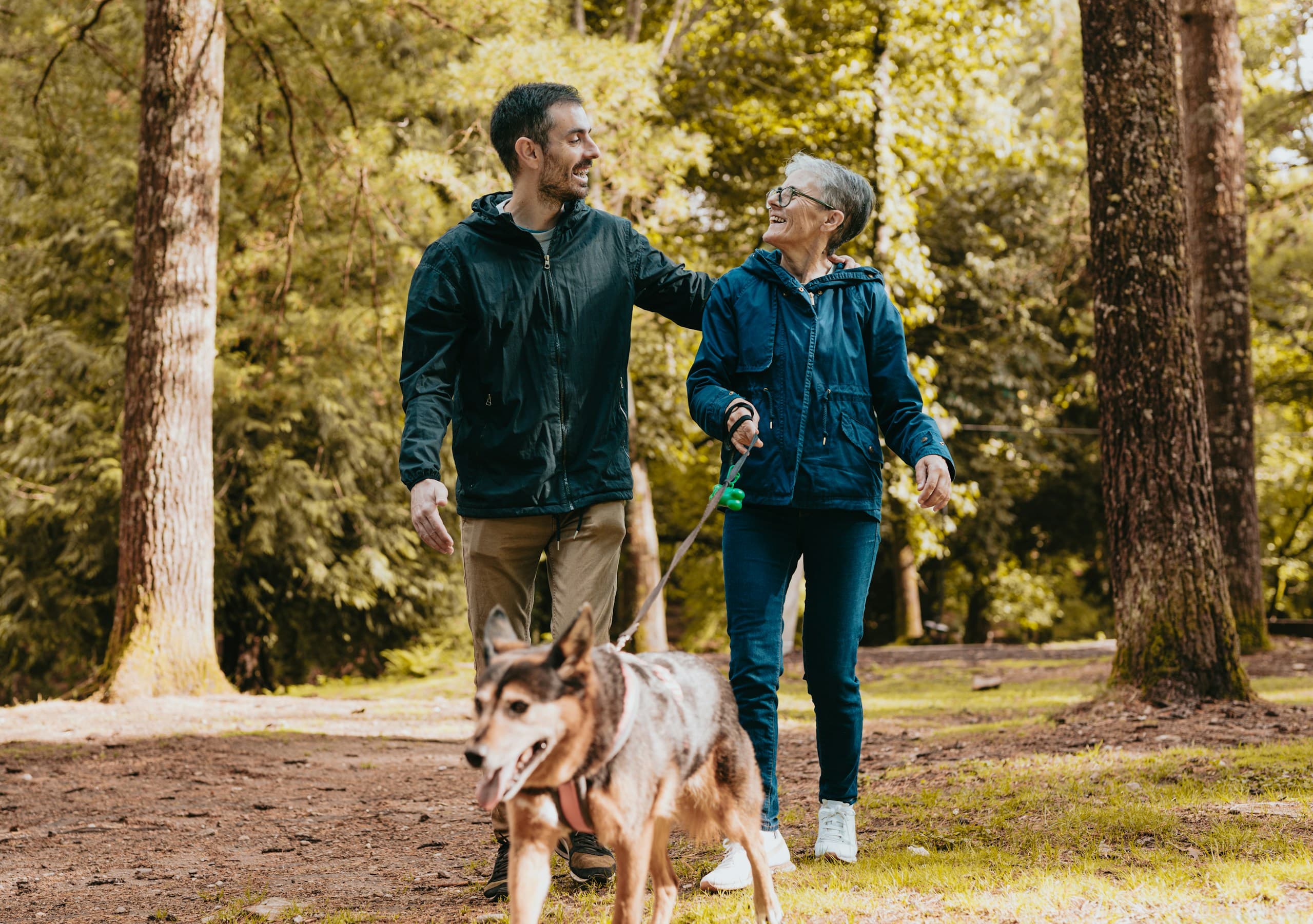A younger man and an older woman walking together through a sunlit forest